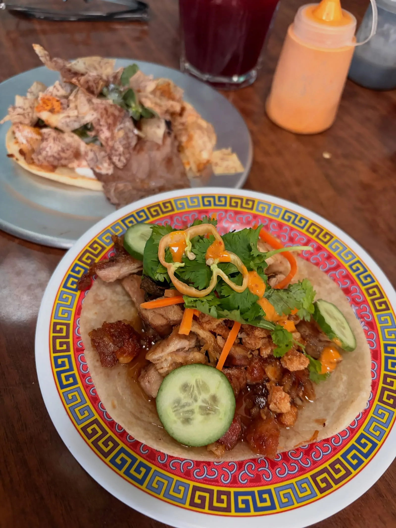 Two tacos on patterned plates, one topped with cucumber, cilantro, and pickled carrots