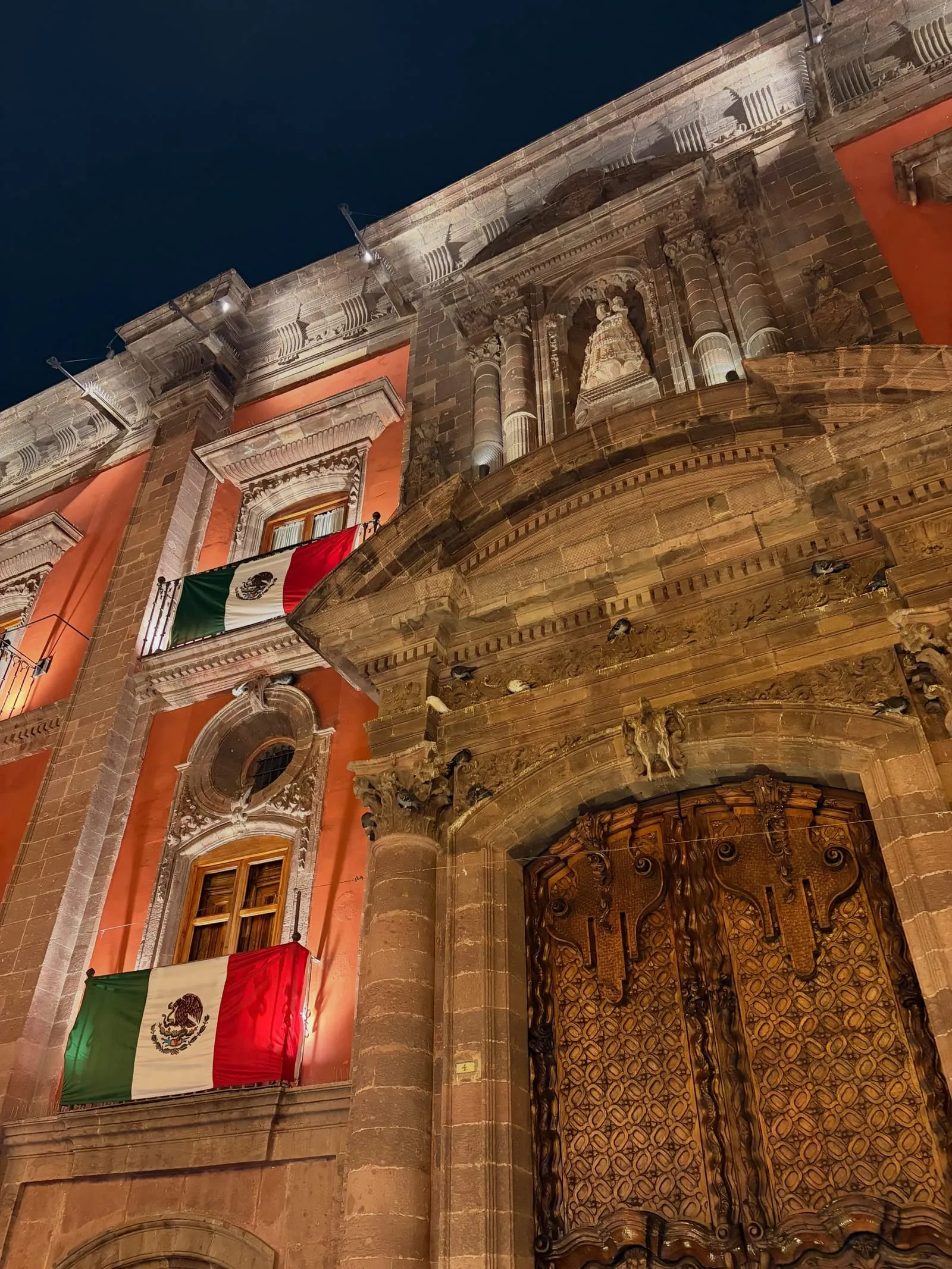 An ornate stone building lit up at night, with Mexican flags hanging from its balconies