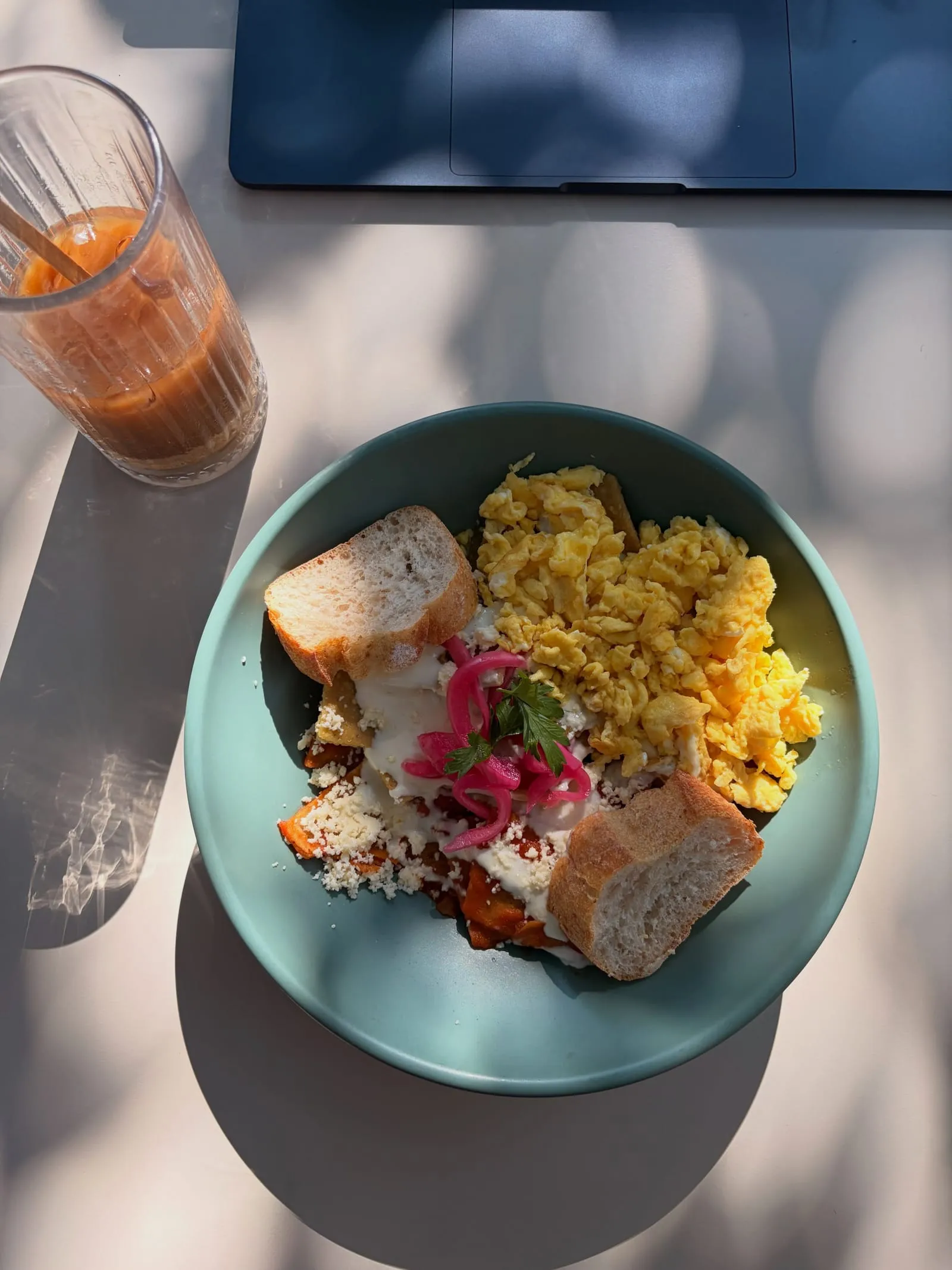 A breakfast bowl of scrambled eggs, chilaquiles, pickled onions, and bread next to an iced coffee