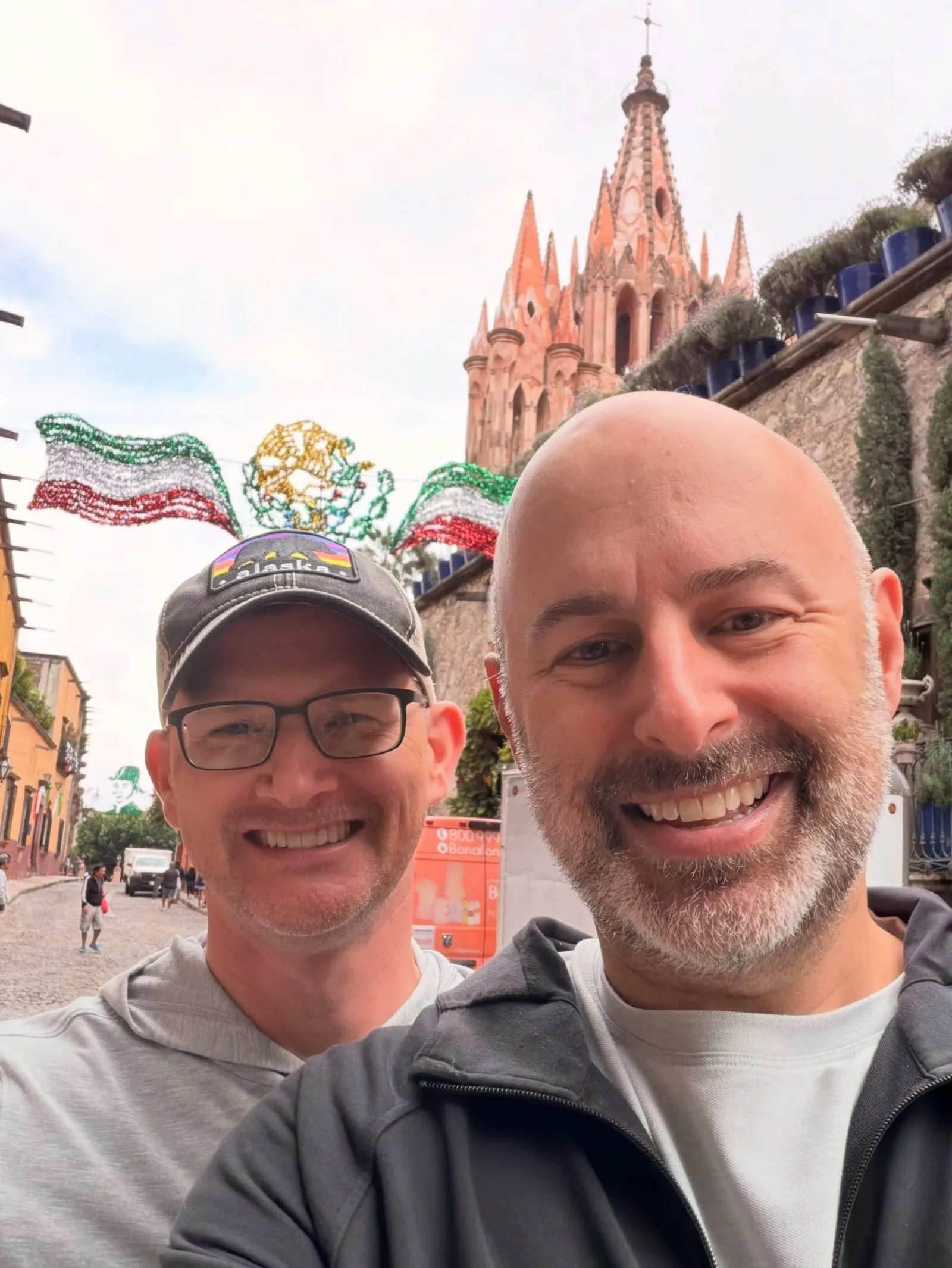 Aaron and a friend taking a selfie on a street in San Miguel de Allende, with the pink spires of the Parroquia behind them