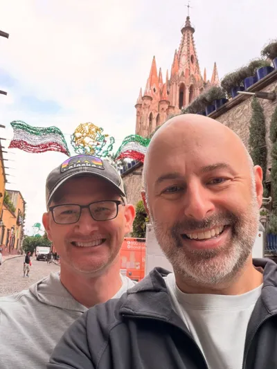 Aaron and a friend taking a selfie on a street in San Miguel de Allende, with the pink spires of the Parroquia behind them